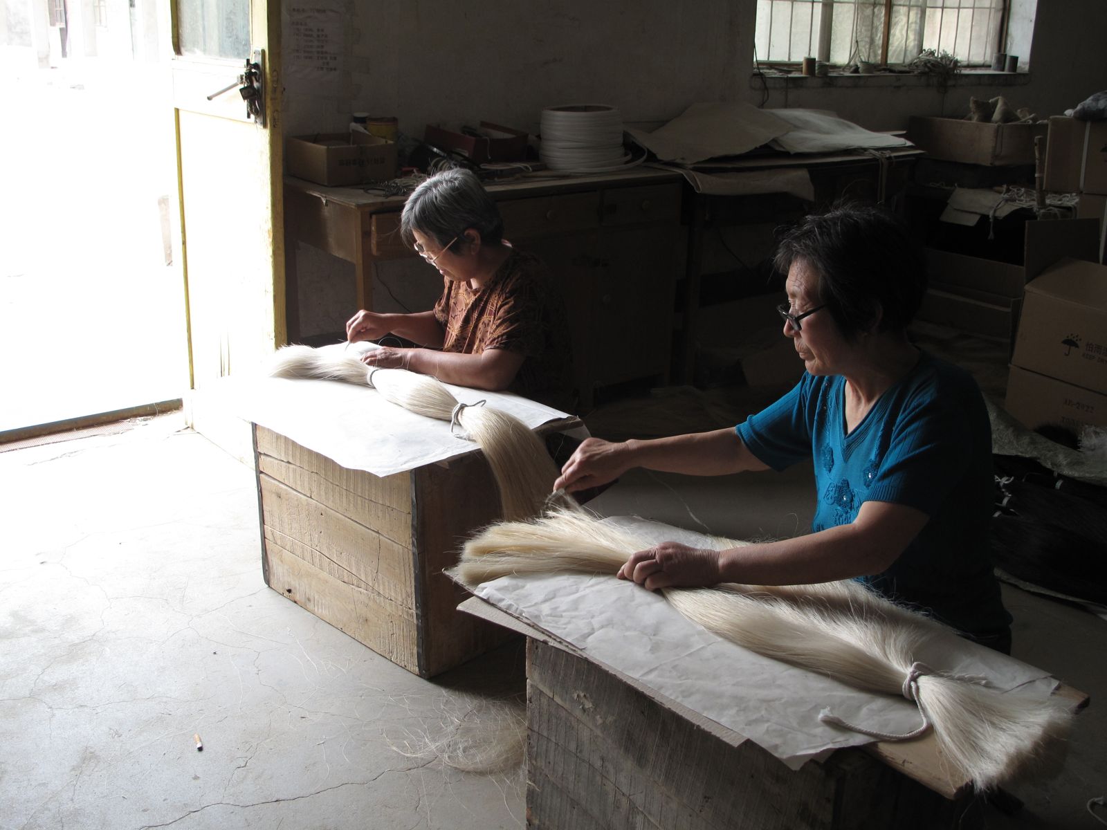 Two women seated at wooden boxes, sorting long hanks of white horsehair by hand