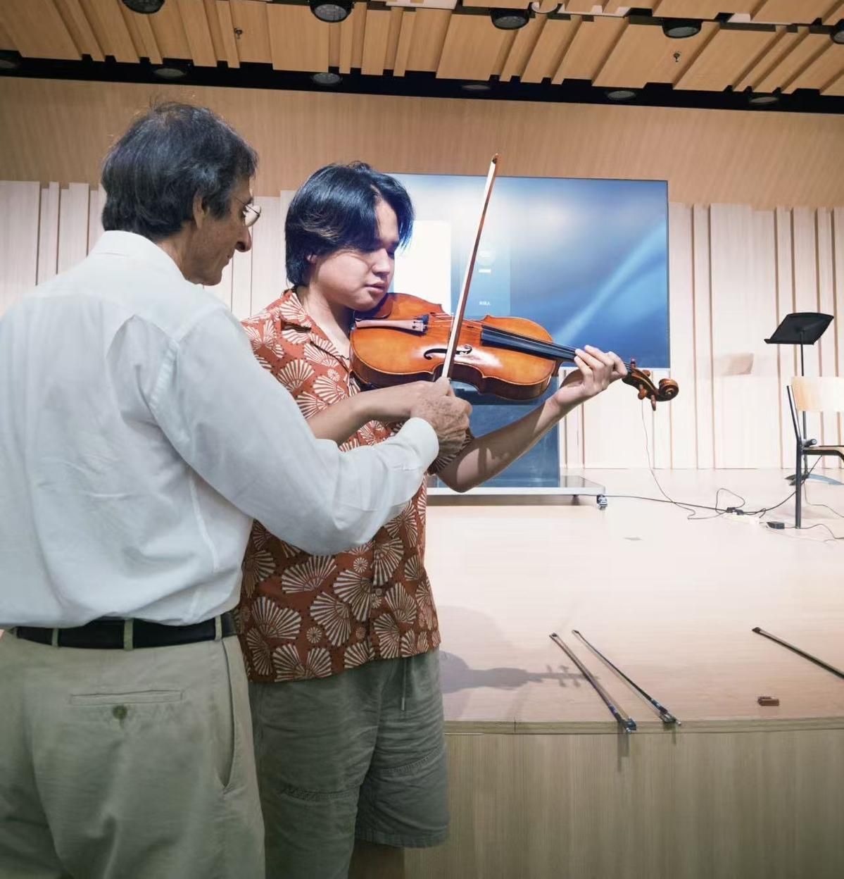 Gilles Saurais standing behind a violin student, guiding the bow arm, with several bows laid out on the stage edge